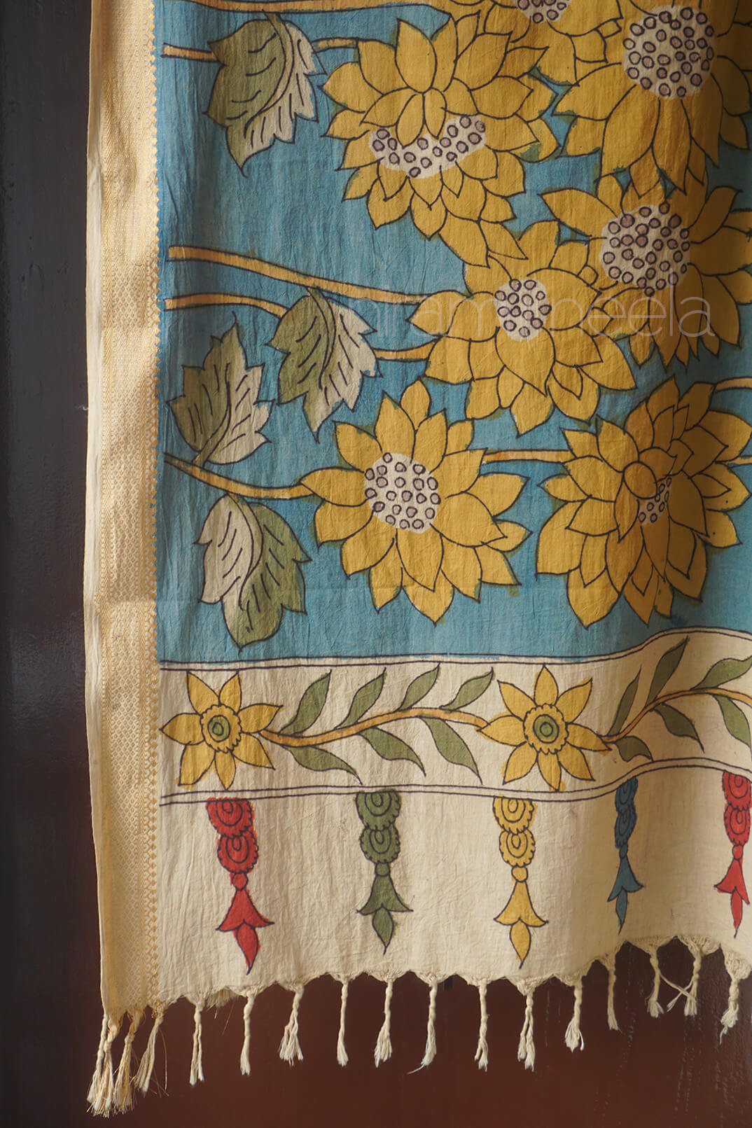 A close-up of a patterned textile, likely a scarf , featuring large yellow sunflower motifs on a turquoise blue background, with a cream-colored lower border, green leaves, and small red and blue decorative motifs. The fabric is draped against a dark brown background.