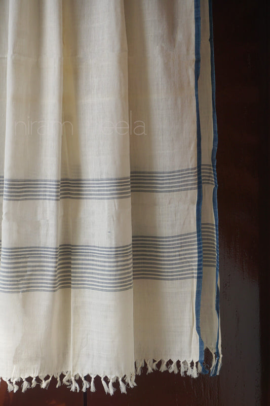 An off-white cotton shawl with thin blue horizontal stripes and a solid blue border, hanging against a dark brown background.