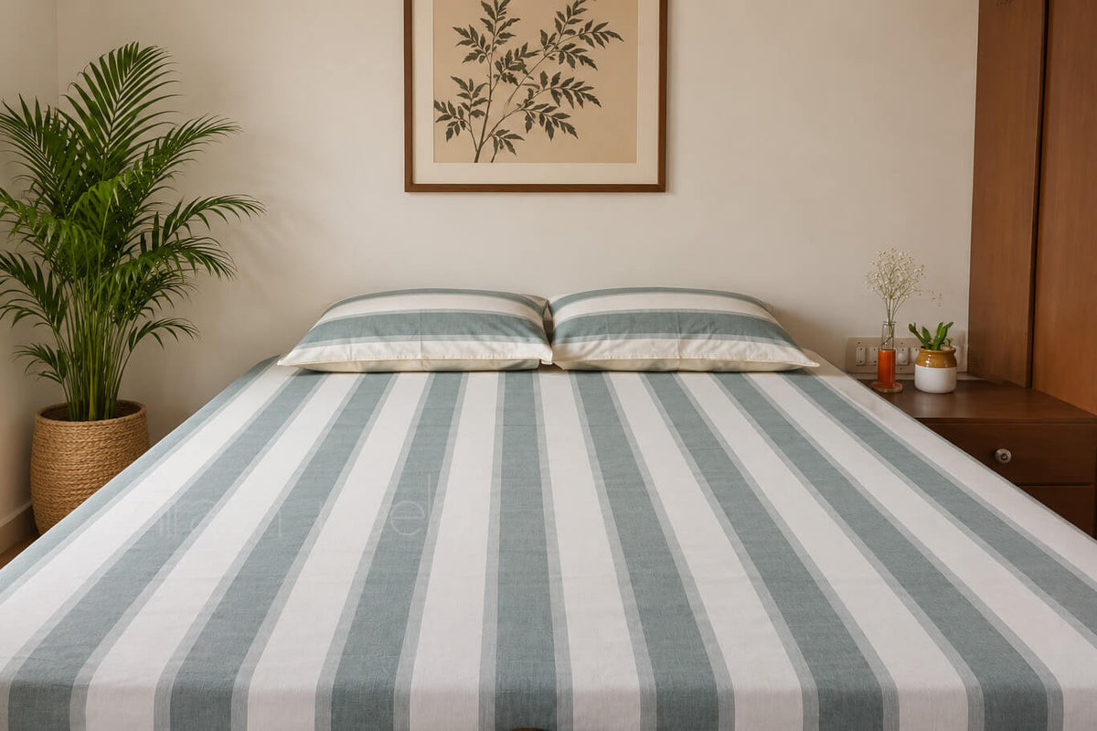 A neatly made bed with a striped sheet and matching pillowcases in muted gray and white, styled in a bedroom with a potted palm, framed wall art, and a wooden nightstand.