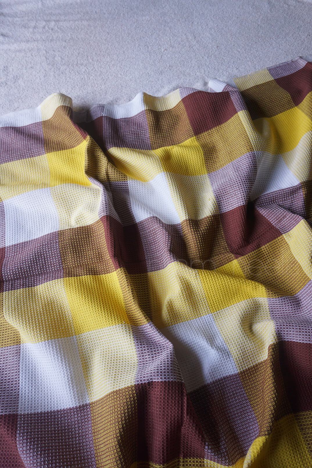 A close-up of a plaid fabric with a textured waffle-weave pattern, featuring yellow, brown, white, and burgundy checks, draped in soft folds.