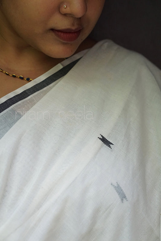 A close-up of a person wearing a white saree with a thin dark border and small black and gray embroidered motifs, photographed against a dark background | Kasavu by Niram Neela