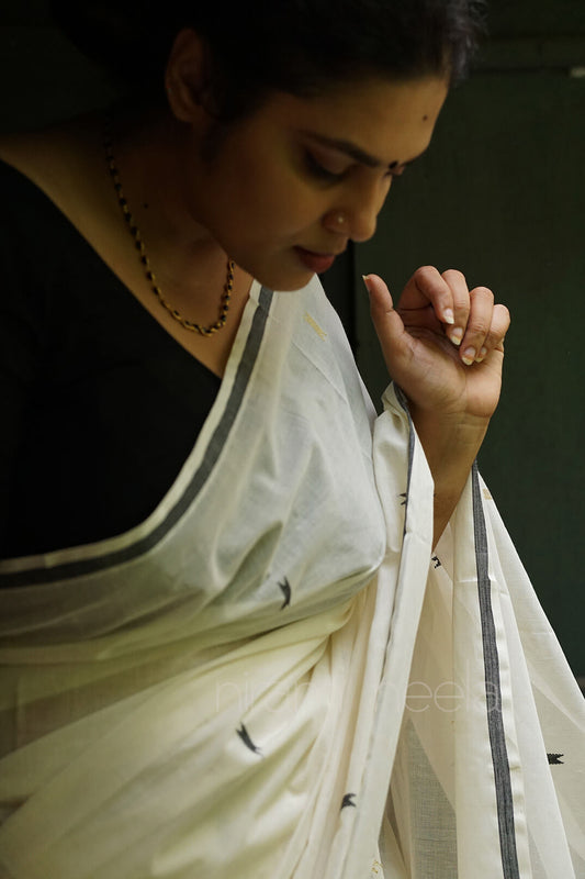 A woman wearing a white sari with black border details and a black blouse, captured in a moody, low-light portrait | Kasavu by Niram Neela