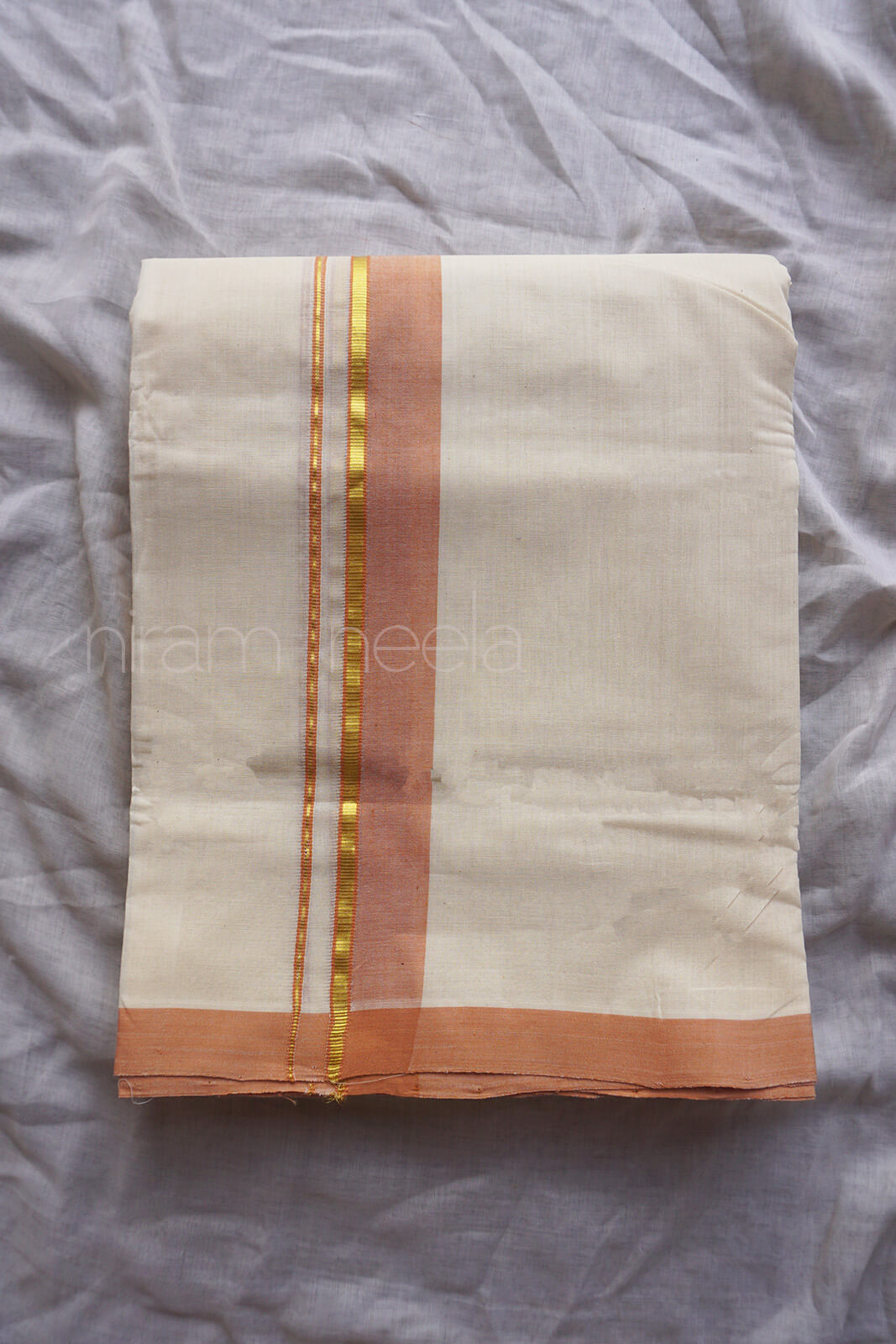 A folded cream-colored cotton dhoti with a peach border and gold zari stripes, laid flat on a wrinkled light gray fabric background.