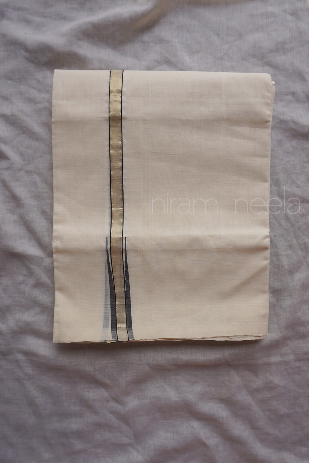 A folded cream-colored cotton dhoti with a vertical metallic gold stripe and thin black accent lines, laid flat on a gray fabric background. A faint watermark 'niram neela' is visible on the right side.