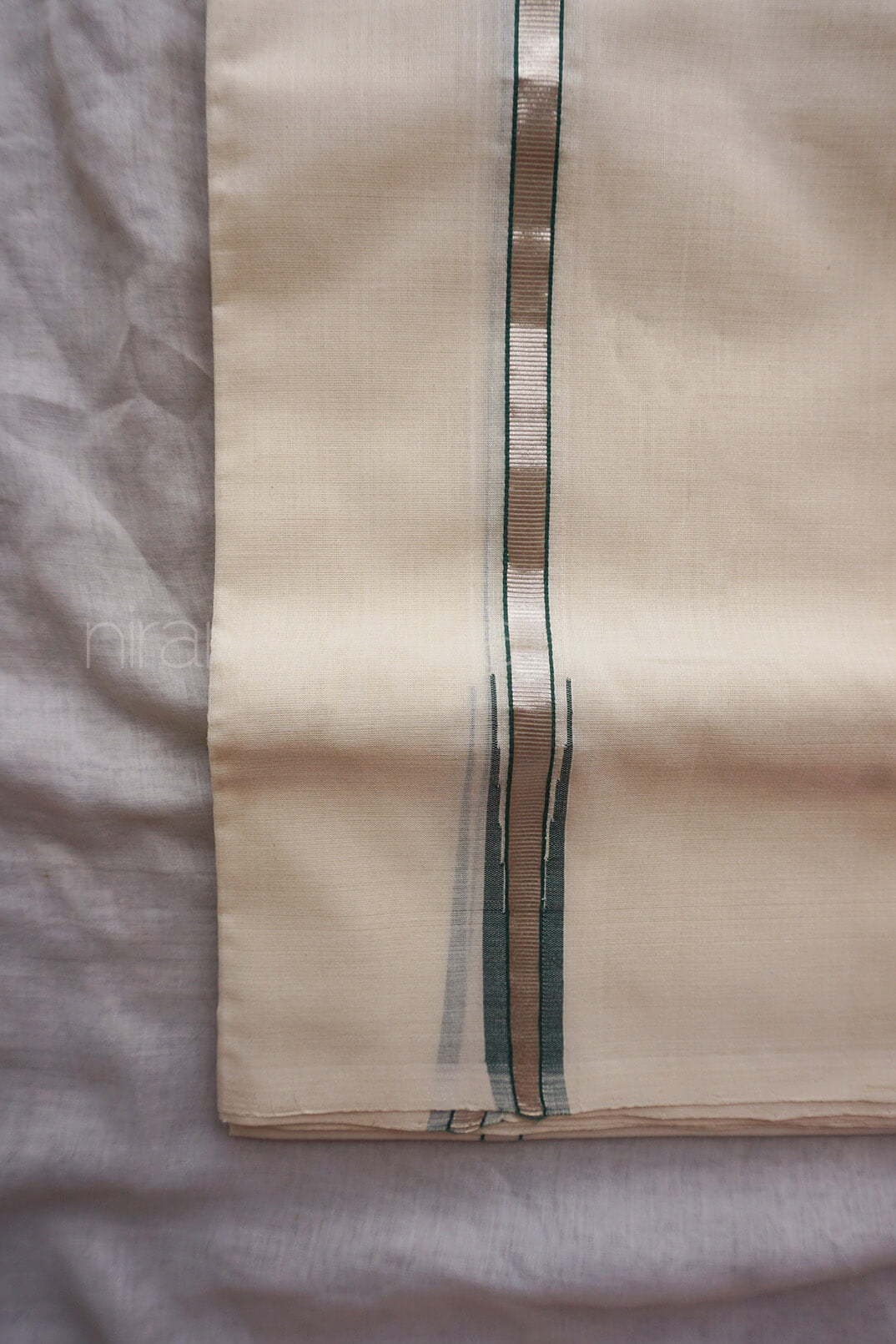 A close-up of a cream-colored dhoti, featuring a central vertical stripe pattern with green, and silver threads, laid flat on a light gray textured fabric background.