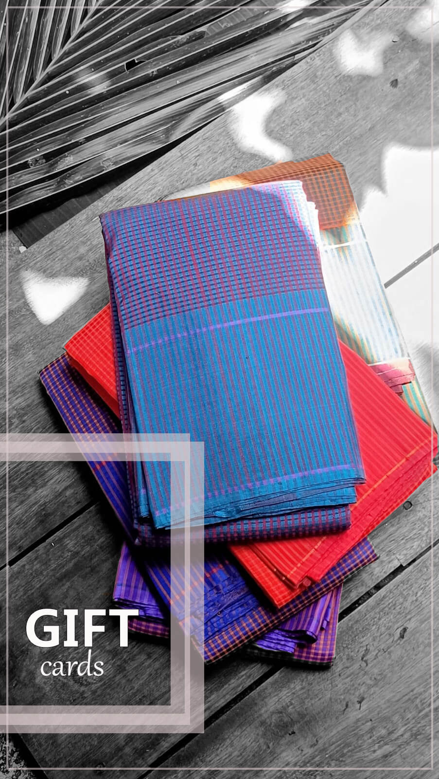 A stack of colorful sarees with a blue, red, and purple plaid pattern, styled on a dark wooden surface with a decorative leaf in the corner. A semi-transparent label in the bottom left reads 'GIFT cards'.