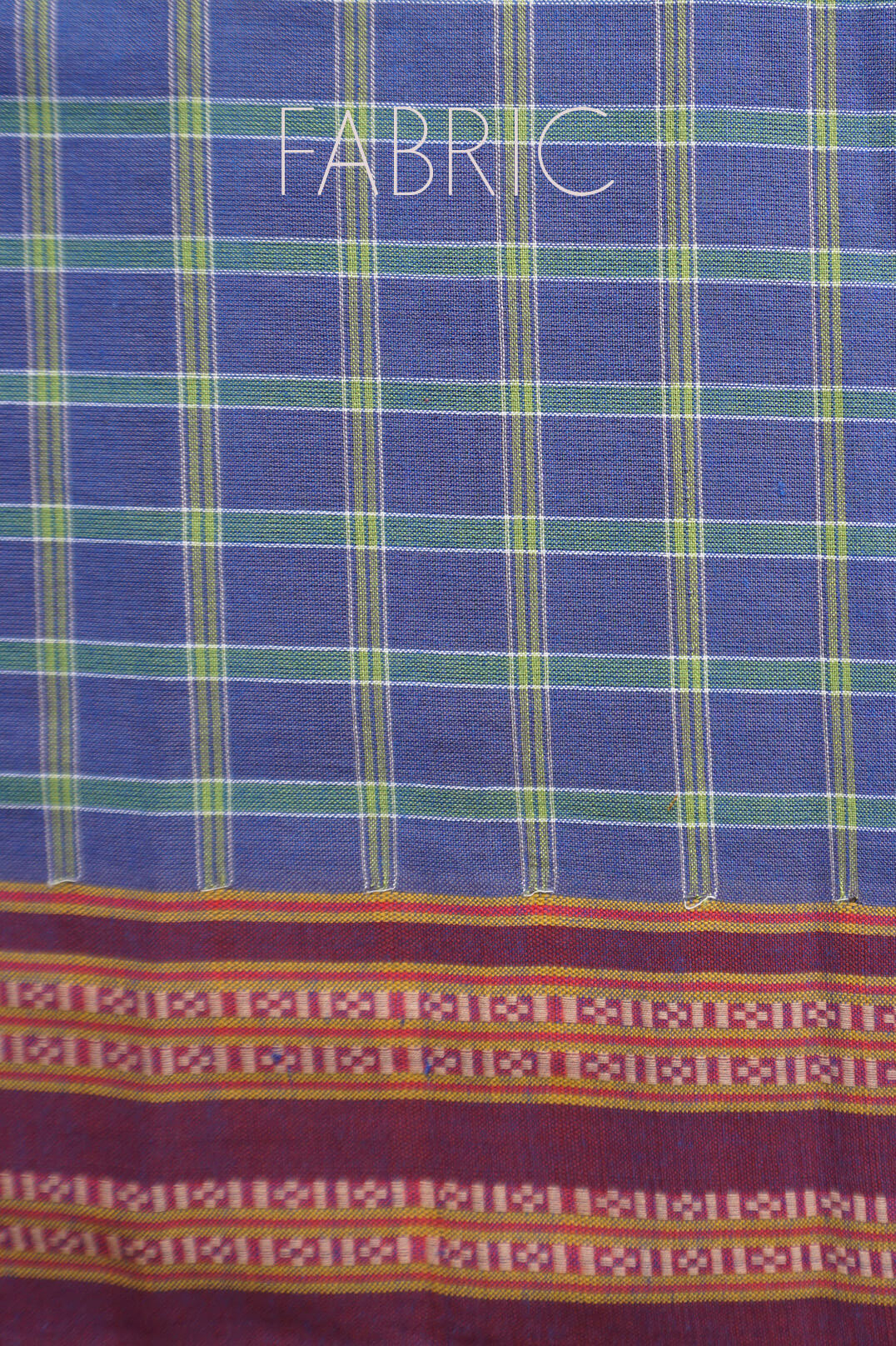 A close-up of a woven Kunbi fabric with a blue base, green and yellow vertical stripes, and a decorative multicolor patterned border at the bottom. The word 'FABRIC' is printed across the top.