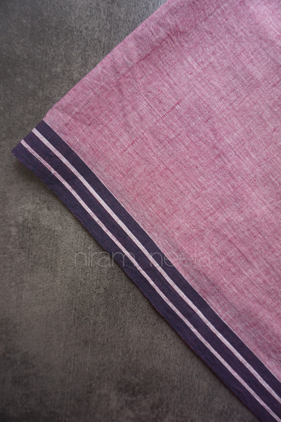 A close-up of an aubergine woven fabric, featuring a black border with thin white stripes, laid diagonally on a textured gray surface.