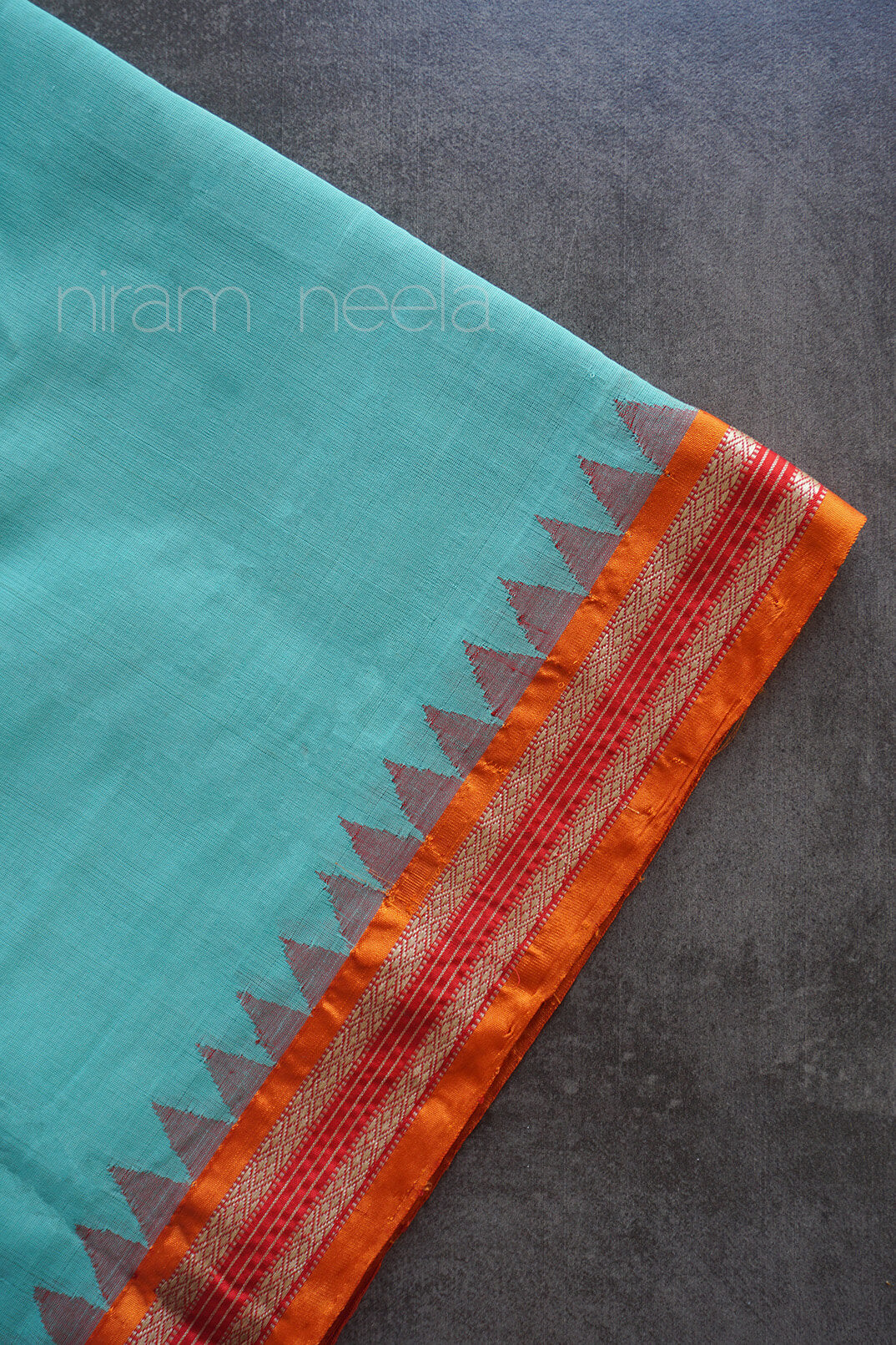 A close-up of a turquoise blue fabric, featuring a decorative border with orange, red, and metallic gold details, laid flat on a dark gray textured surface. The brand name 'niram neela' is printed in white text on the upper left.