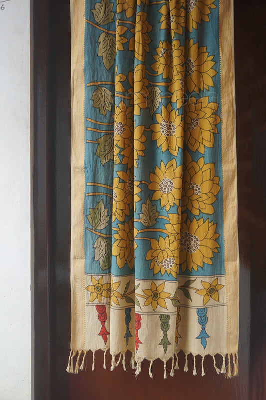A handpainted Kalamkari cotton duppatta  hanging in front of a dark brown door. The fabric features a teal blue background with large yellow floral motifs, a beige border, and small colorful handpainted details near the bottom fringe.