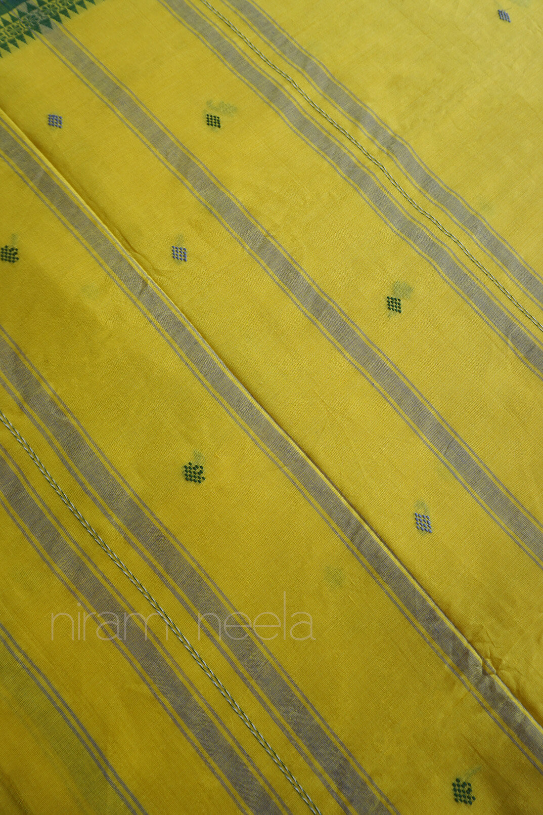 A close-up of a yellow saree pallu with diagonal gray stripes and small green embroidered motifs, showing the textile texture and pattern.
