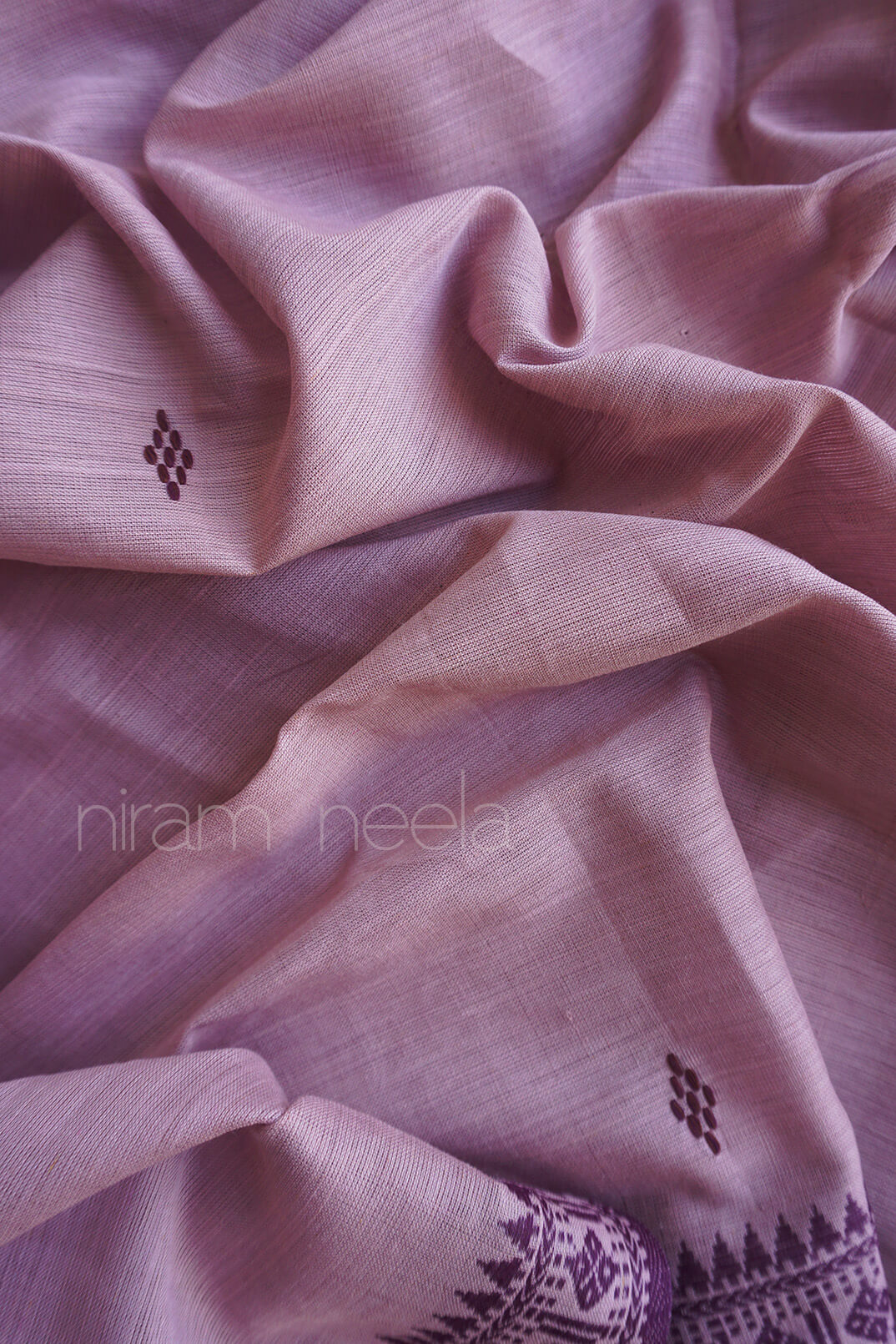A close-up of draped dusty purple fabric with small dark embroidered motifs and a patterned border visible at the bottom edge. The watermark 'niram neela' appears across the center.