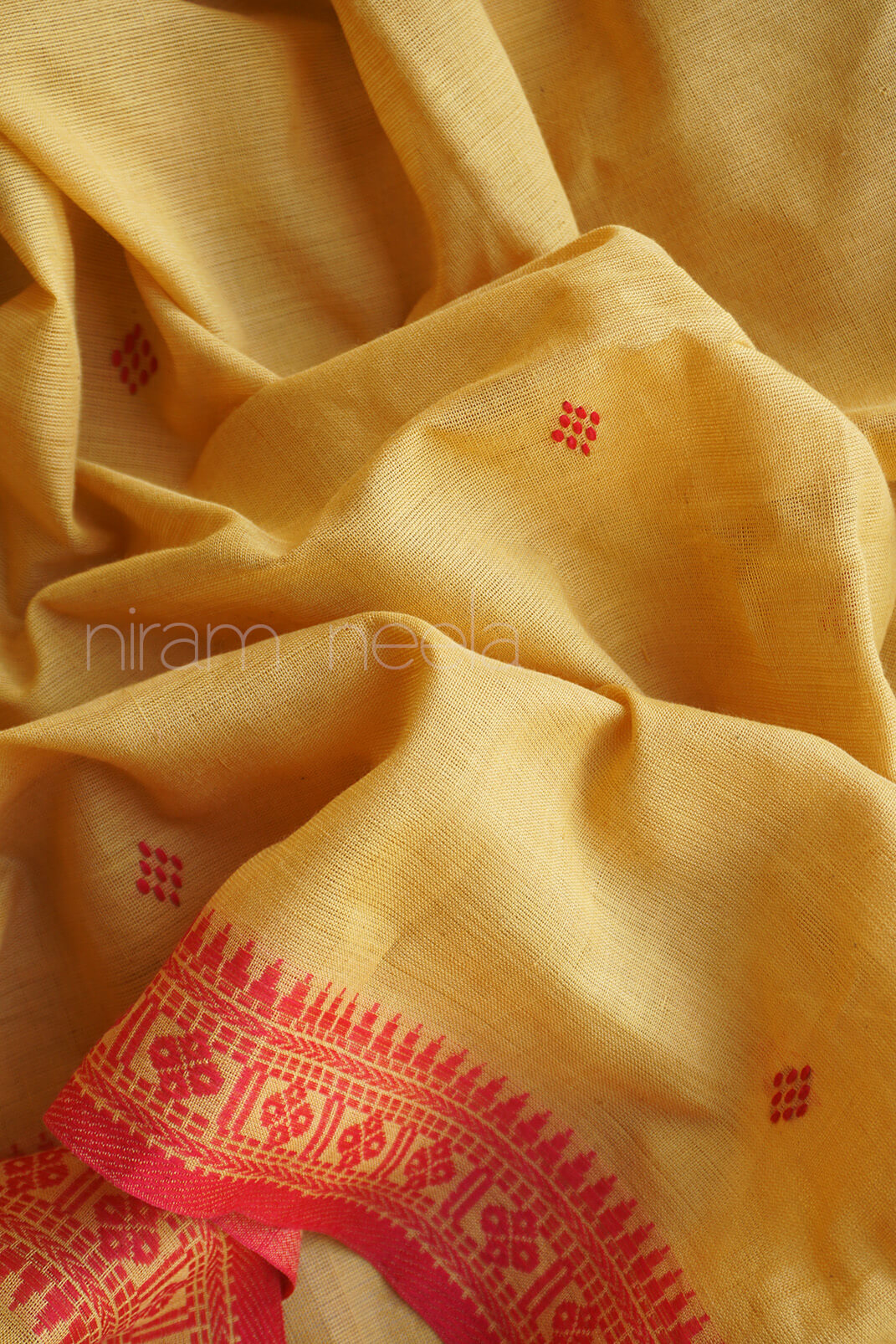 A close-up of draped mustard yellow saree with small red embroidered motifs and a decorative red border along one edge. A faint 'niram neela' watermark is visible across the center.