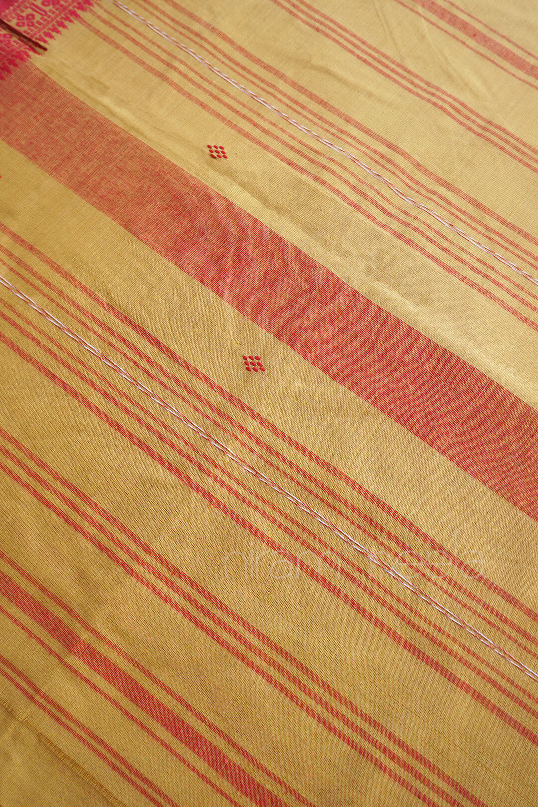 A close-up of a saree, featuring a warm beige base with diagonal coral-red stripes and small red diamond motifs with visible 'Niram Neela' branding.