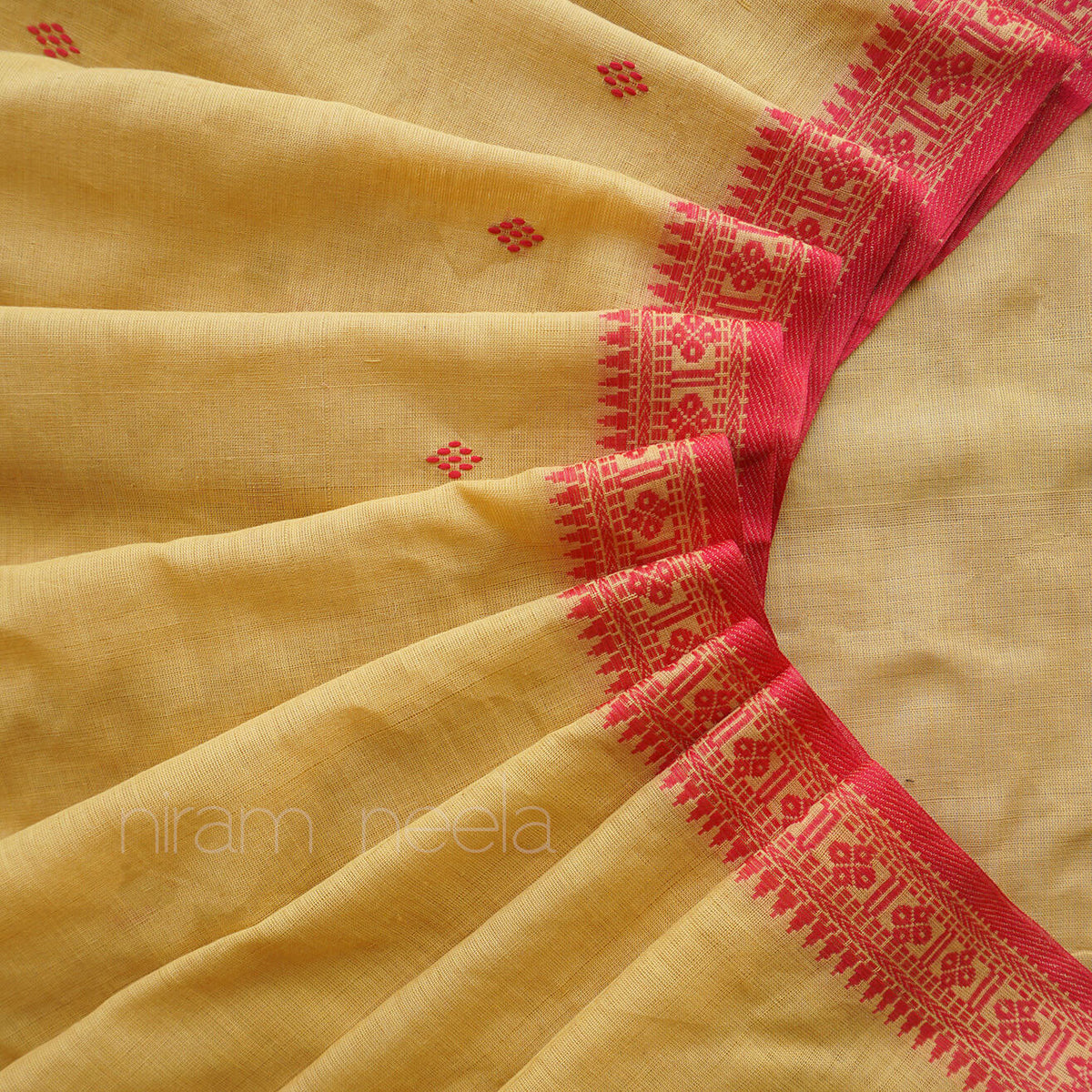 A close-up of a pale yellow saree, with a vibrant red border featuring intricate geometric and floral patterns. Small red diamond motifs are scattered across the main body of the fabric.