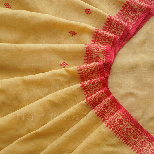 A close-up of a pale yellow saree, with a vibrant red border featuring intricate geometric and floral patterns. Small red diamond motifs are scattered across the main body of the fabric.
