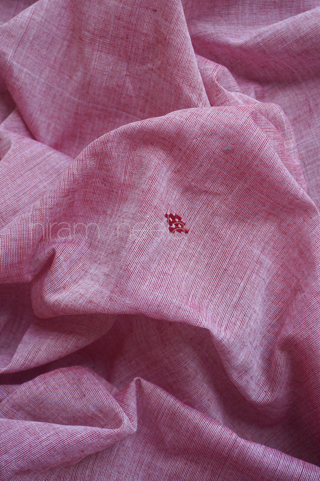 A close-up of draped pink woven fabric with a small red embroidered motif near the center with visible 'Niram Neela' branding