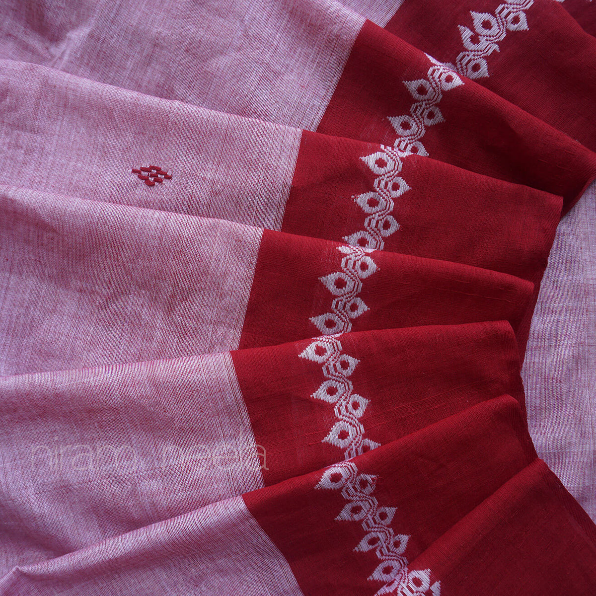 A close-up of a draped sareee, featuring a greyish pink body with a deep red border decorated with white geometric embroidery with visible 'Niram Neela' branding