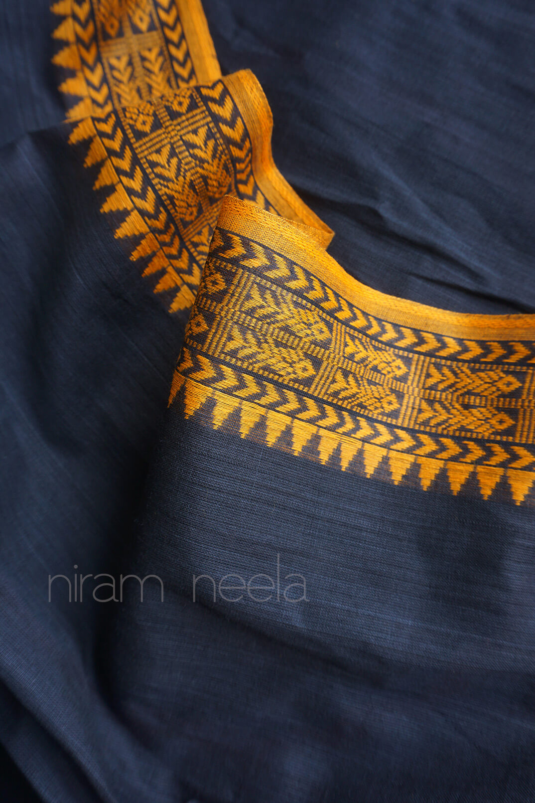 A close-up of a dark gray saree with a wide yellow woven border featuring geometric and tribal patterns. The brand name 'niram neela' is watermarked in the lower left corner.