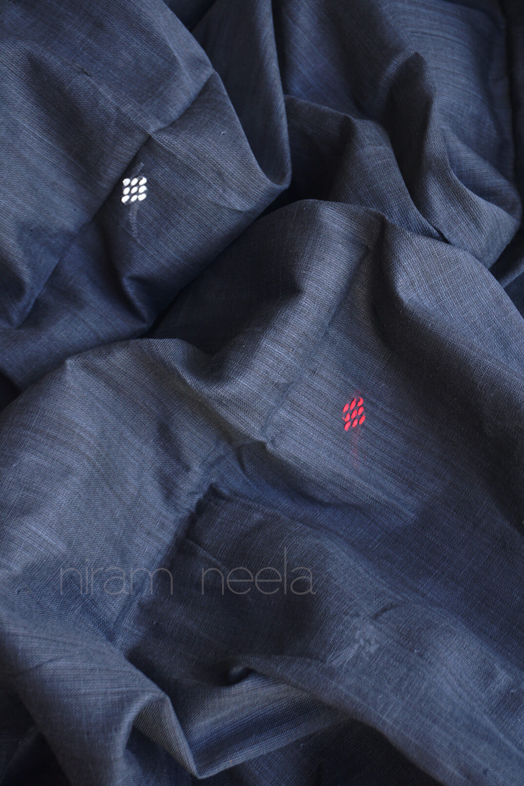 A close-up of draped dark gray fabric with a subtle woven texture, featuring two small woven geometric motifs in white and red, and a faint watermark reading 'niram neela' across the lower portion.