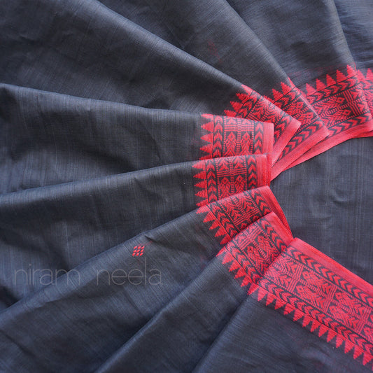 A close-up of a dark charcoal gray saree, draped in a fan-like arrangement  with visible ' Niram Neela' branding. The fabric features a vibrant red border with black traditional geometric and floral patterns.