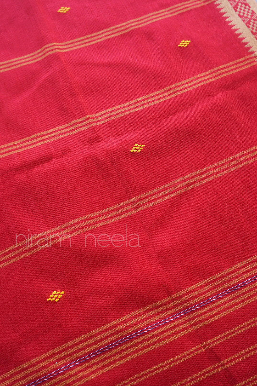 A close-up of a red saree, featuring horizontal dust brown stripes and small yellow geometric motifs. A purple braided border is visible along the bottom right edge with 'Niram Neela' branding visible