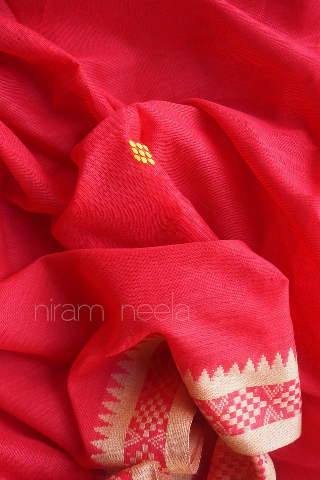 A close-up of a red saree, draped to show its texture and a decorative border with a small yellow woven motif and a dust brown and red patterned edge. The watermark 'niram neela' is visible on the lower left.