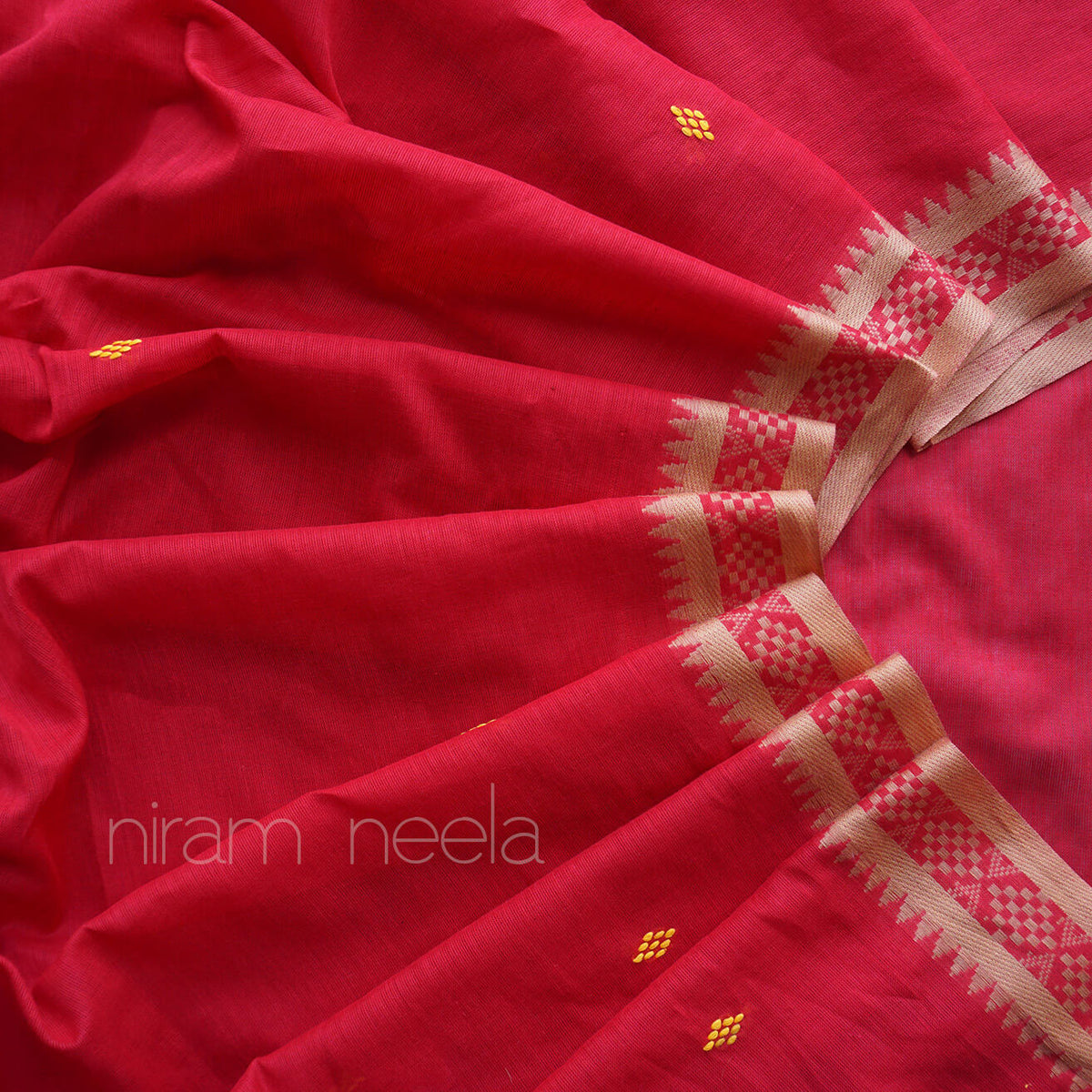 A close-up of a red saree, featuring a dust brown and red checkered border with small yellow woven motifs. The brand name 'niram neela' is printed in white text in the lower-left corner.