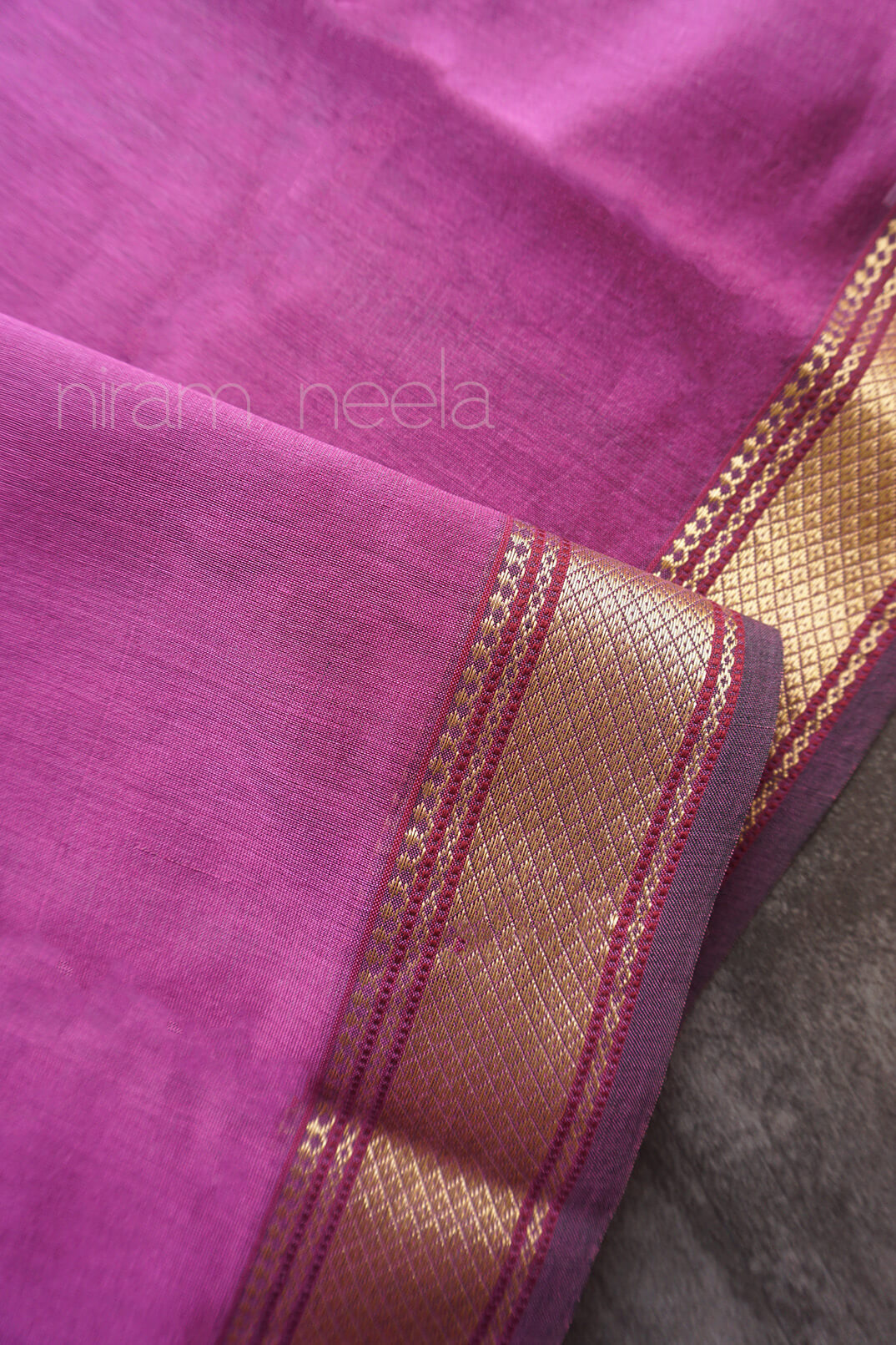 A close-up of a purple silk cotton fabric, featuring a prominent metallic gold border with woven patterns and thin red accent stripes. The brand name 'niram neela' is faintly watermarked on the upper left.