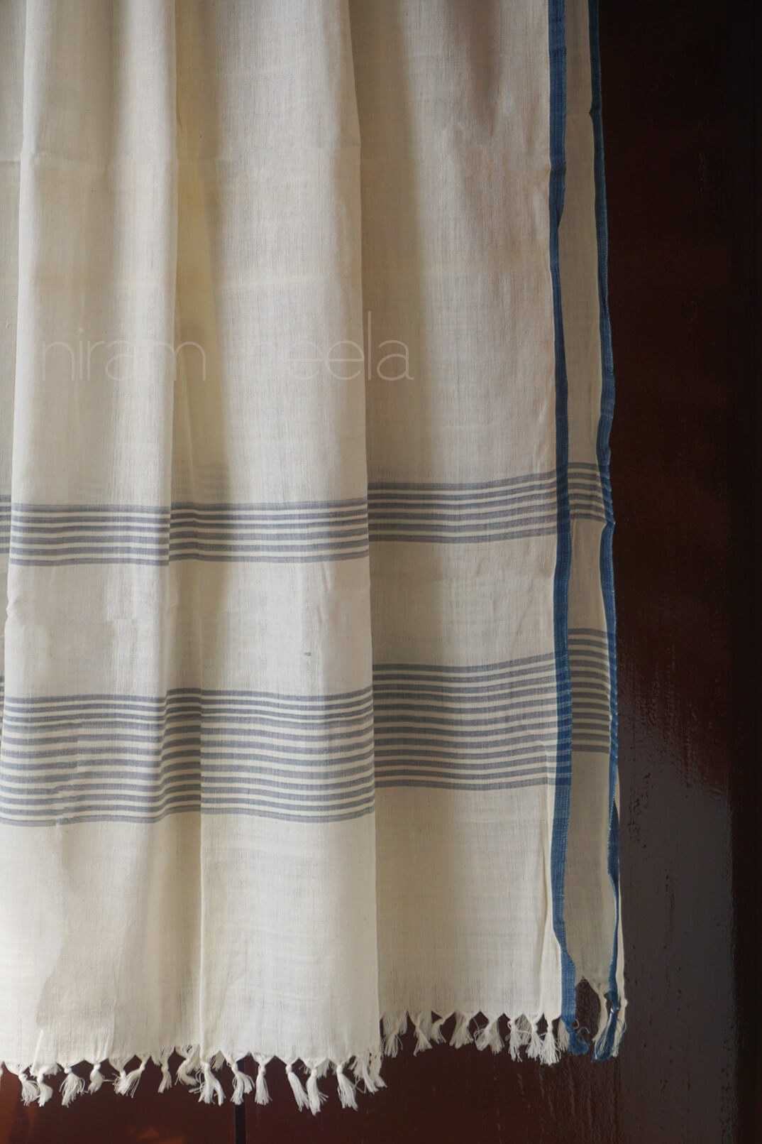 An off-white cotton shawl with thin blue horizontal stripes and a solid blue border, hanging against a dark brown background.