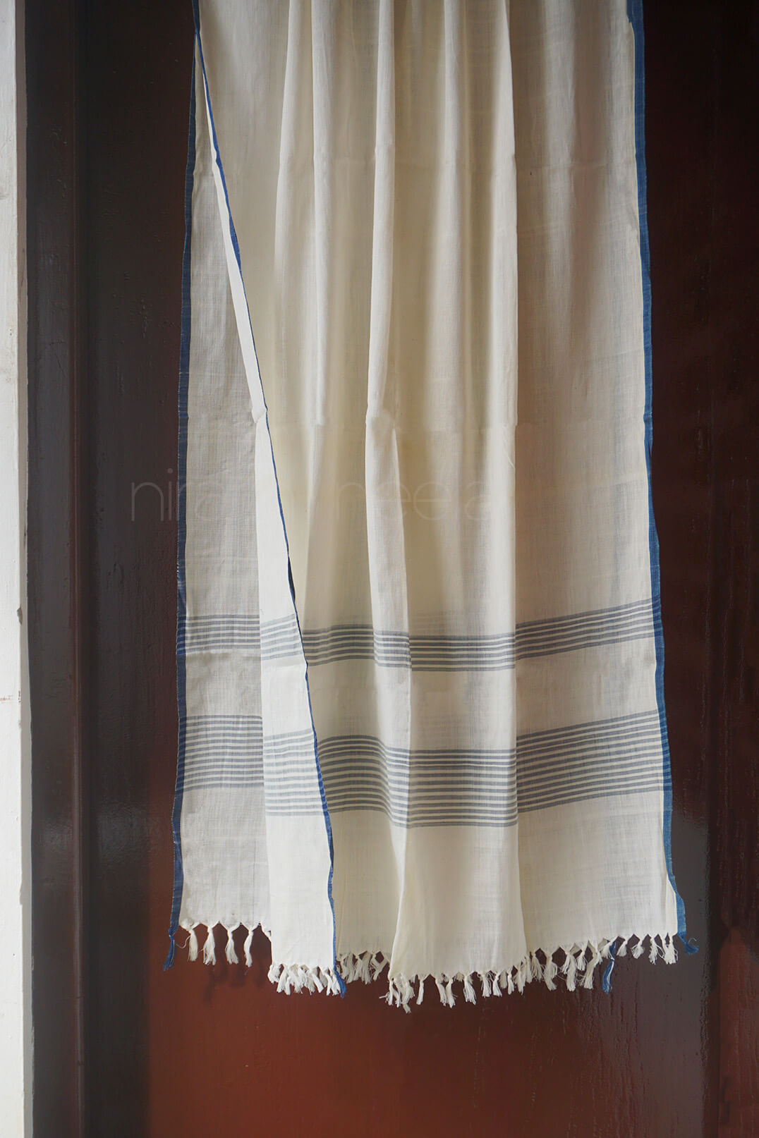 An off-white cotton shawl with thin blue stripes and fringed edges, hanging against a dark brown wooden door.