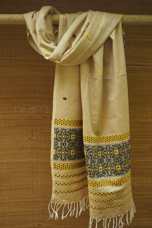 Beige scarf with black and yellow patterns draped over a wooden rod on a brown background.