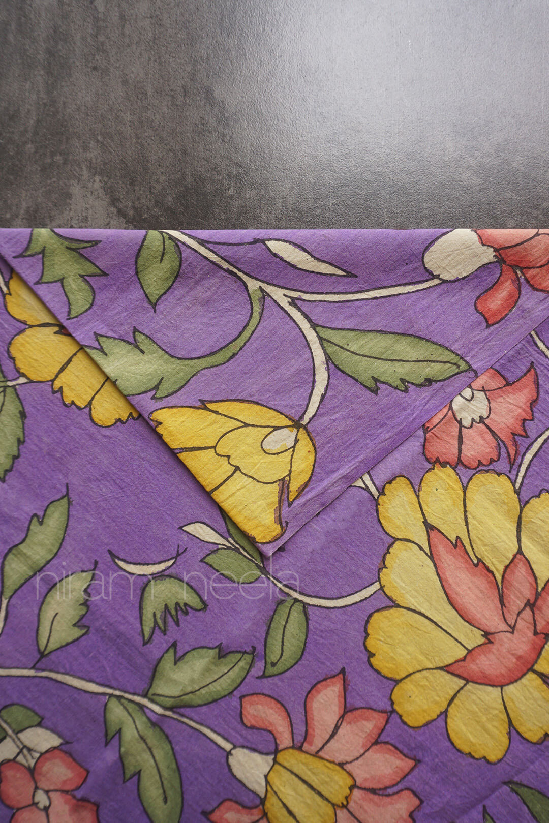 A close-up of a violet fabric with a vintage floral pattern featuring yellow, pink, and green flowers and leaves, folded over on a dark gray surface.