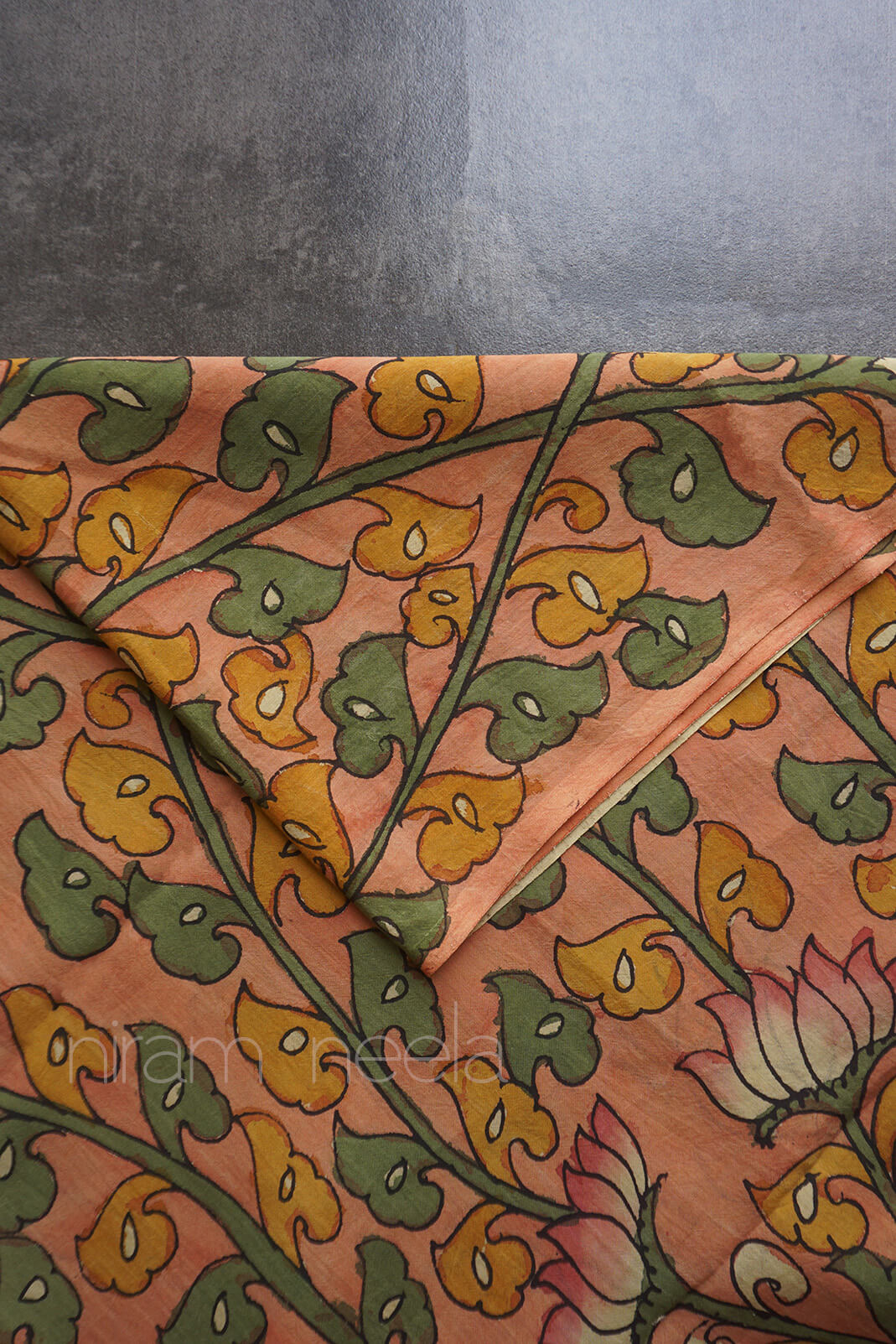 A close-up of a peach-colored fabric with a hand-painted floral design in green and mustard yellow, featuring a lotus flower motif.