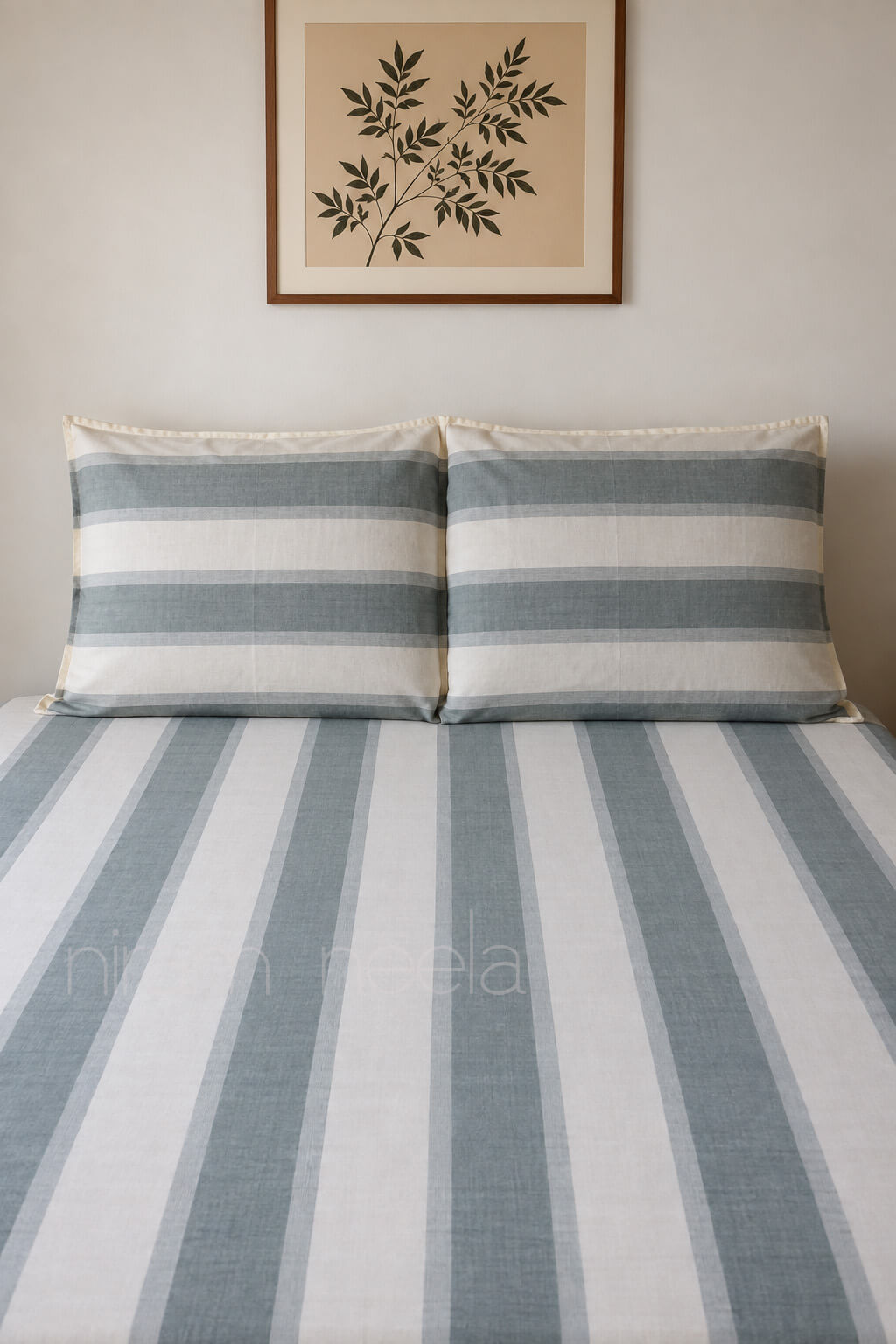 A bed dressed in a striped bedding set with two matching pillowcases, featuring vertical stripes in muted blue-gray and off-white. The bed is styled against a neutral beige wall with a framed botanical print hanging above.