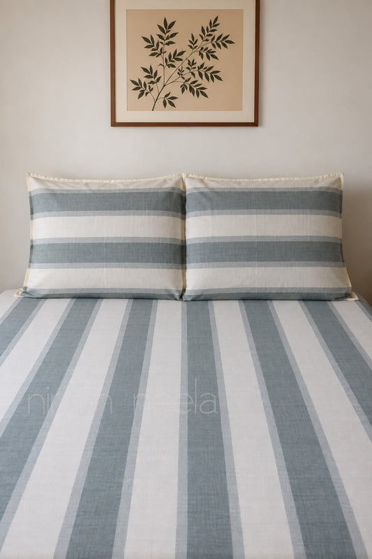 A bed dressed in a striped bedding set with two matching pillowcases, featuring vertical stripes in muted blue-gray and off-white. The bed is styled against a neutral beige wall with a framed botanical print hanging above.