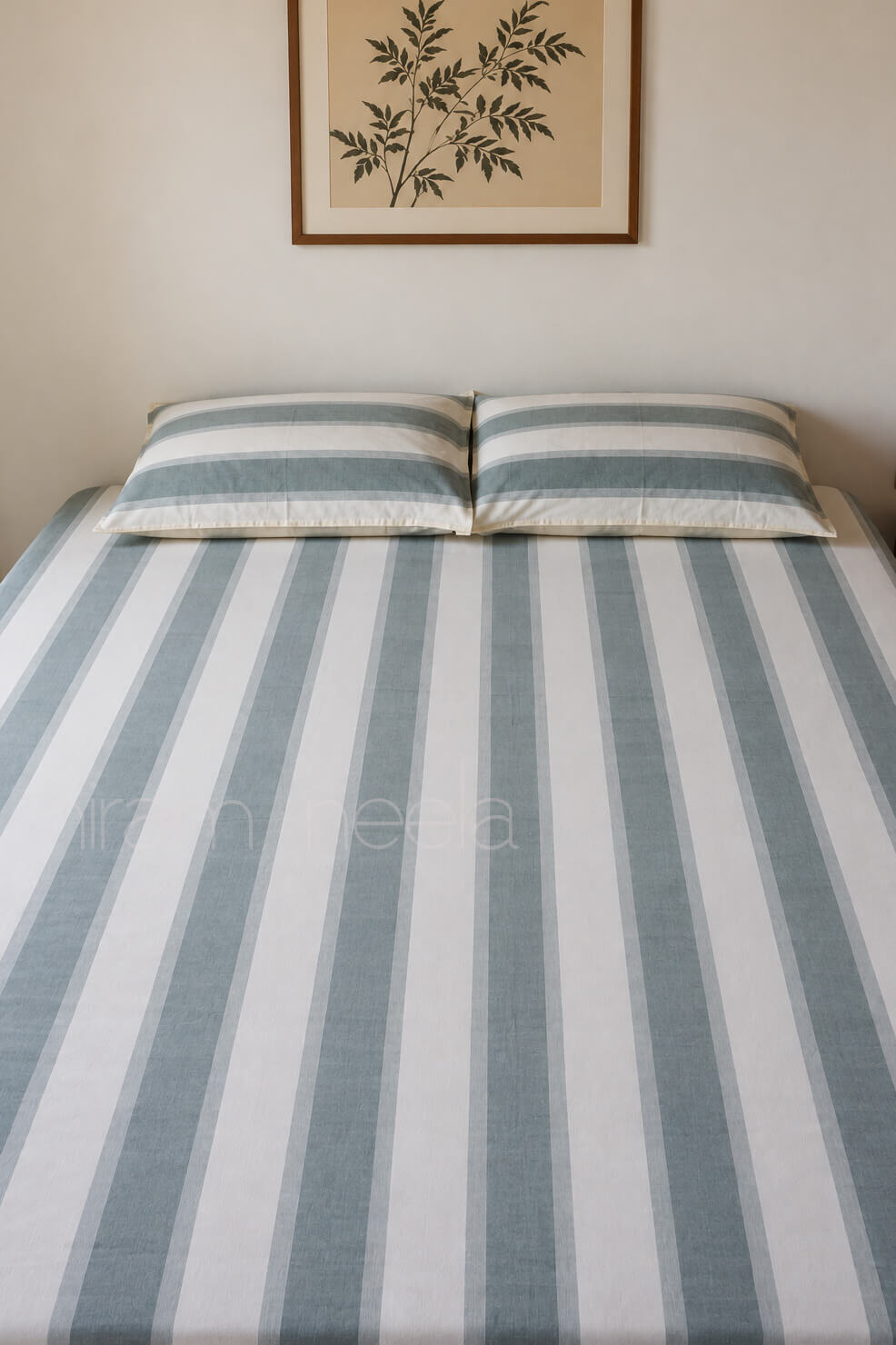 A bed dressed in a striped sheet and matching pillowcases with vertical bands of muted gray and off-white, styled in a minimalist bedroom setting with a framed botanical print on the wall.