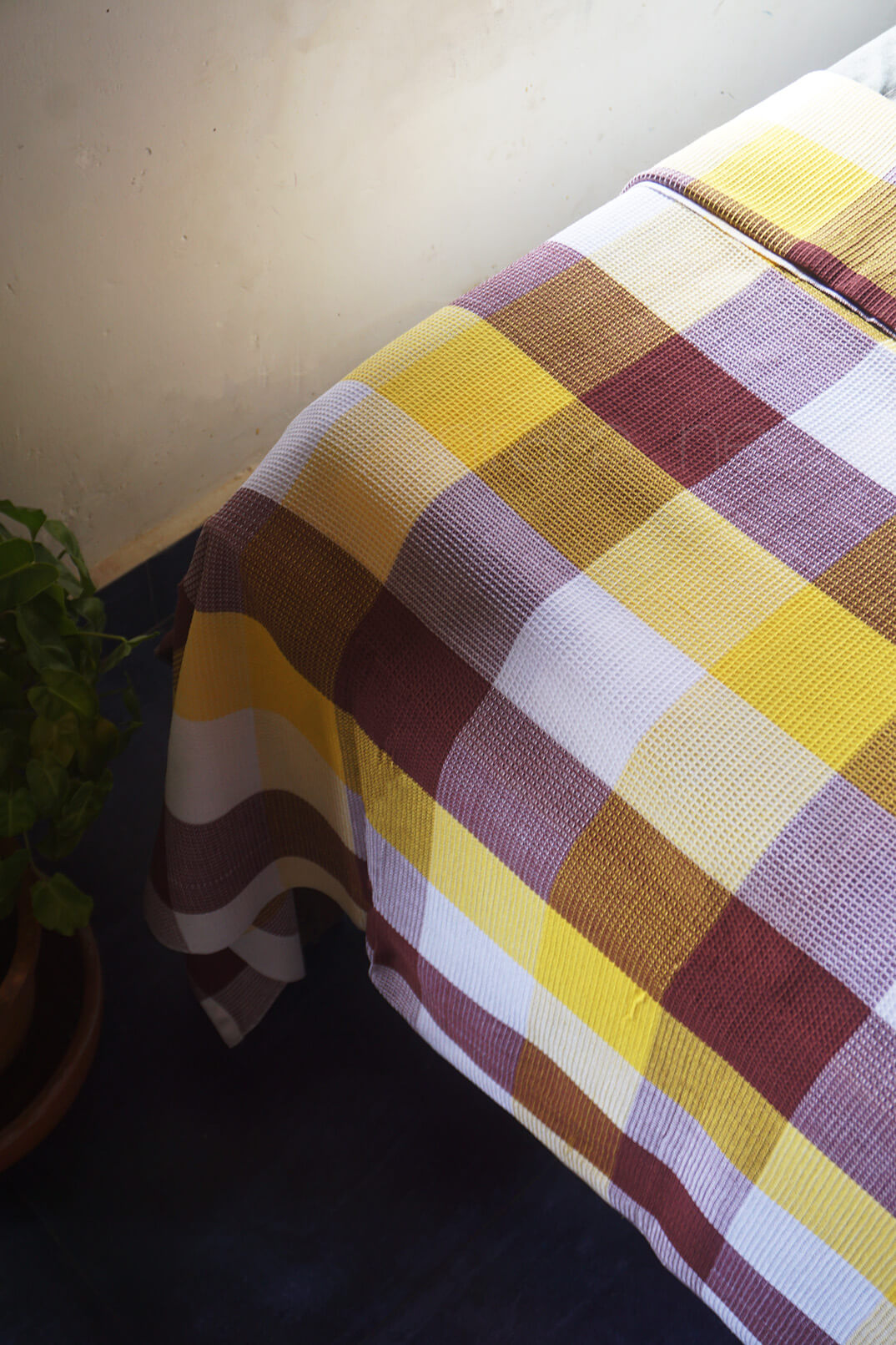 A close-up of a textured plaid tablecloth draped over a table corner, featuring a checkered pattern of yellow, burgundy, white, and brown, with a dark blue floor and a potted plant visible on the left.