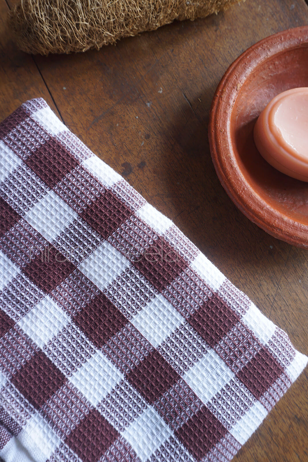 Checkered bath towel, loofah, and small ceramic dish with soap on a wooden surface | Bed and bath by Niram Neela