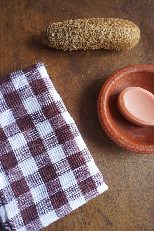 Checkered bath towel, loofah, and small ceramic dish with soap on a wooden surface | Bed and bath by Niram Neela