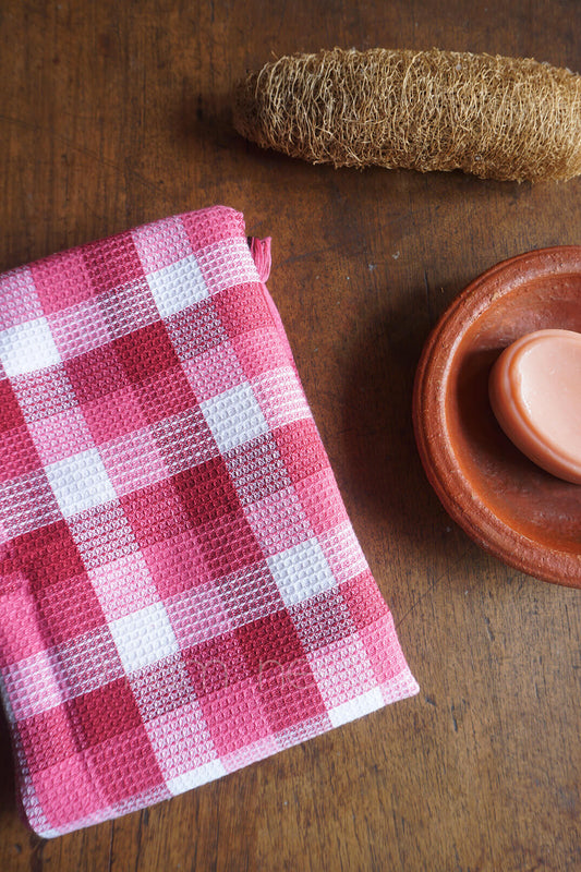 Pink checkered bath towel on a wooden surface with a loofah and small bowl | Bath linen by Niram Neela