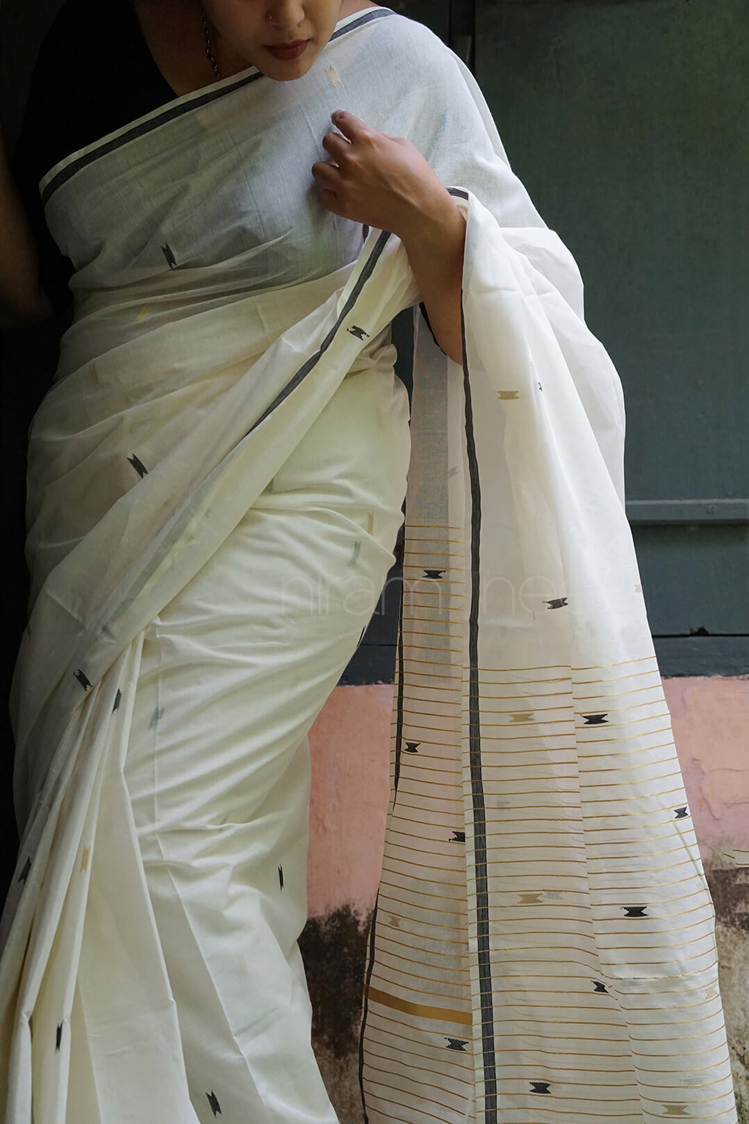A woman wearing a white cotton saree with a black border and small black woven motifs on the pallu, standing against a muted background.