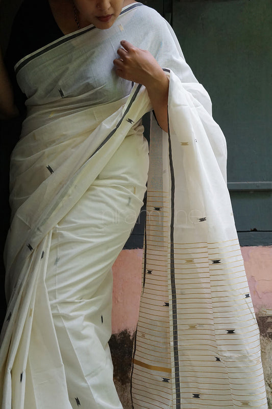 A woman wearing a white cotton saree with a black border and small black woven motifs on the pallu, standing against a muted background.