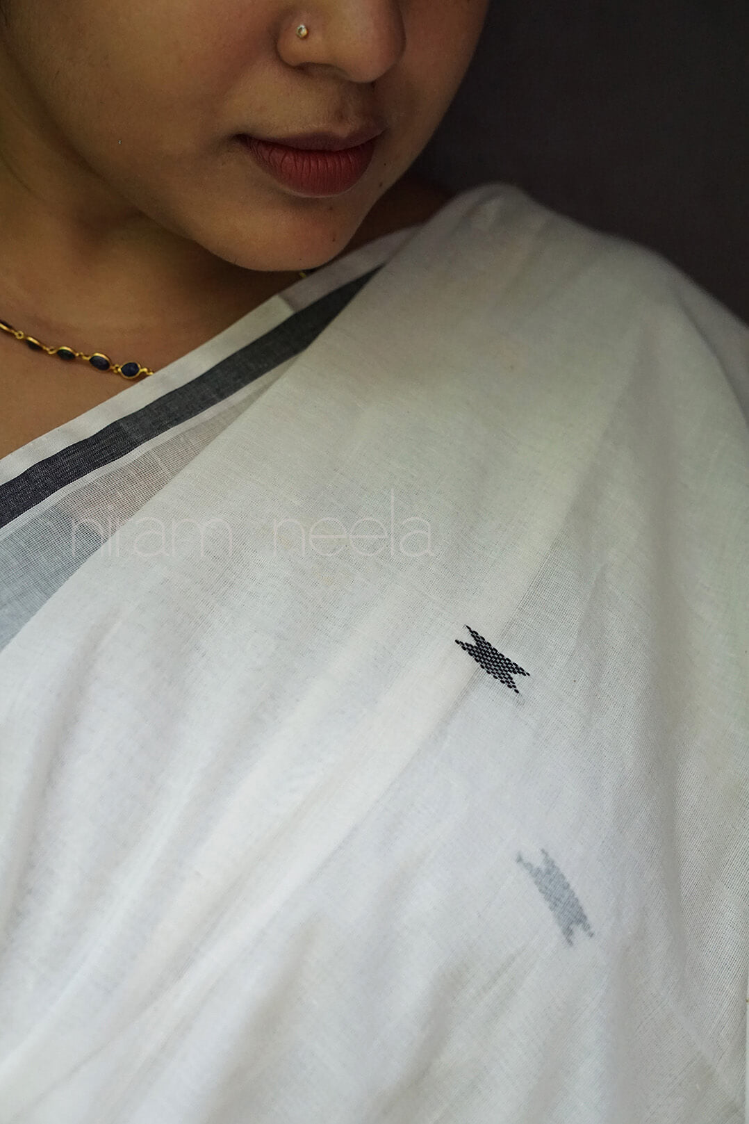 A close-up of a person wearing a white saree with a thin dark border and small black and gray embroidered motifs, photographed against a dark background | Kasavu by Niram Neela