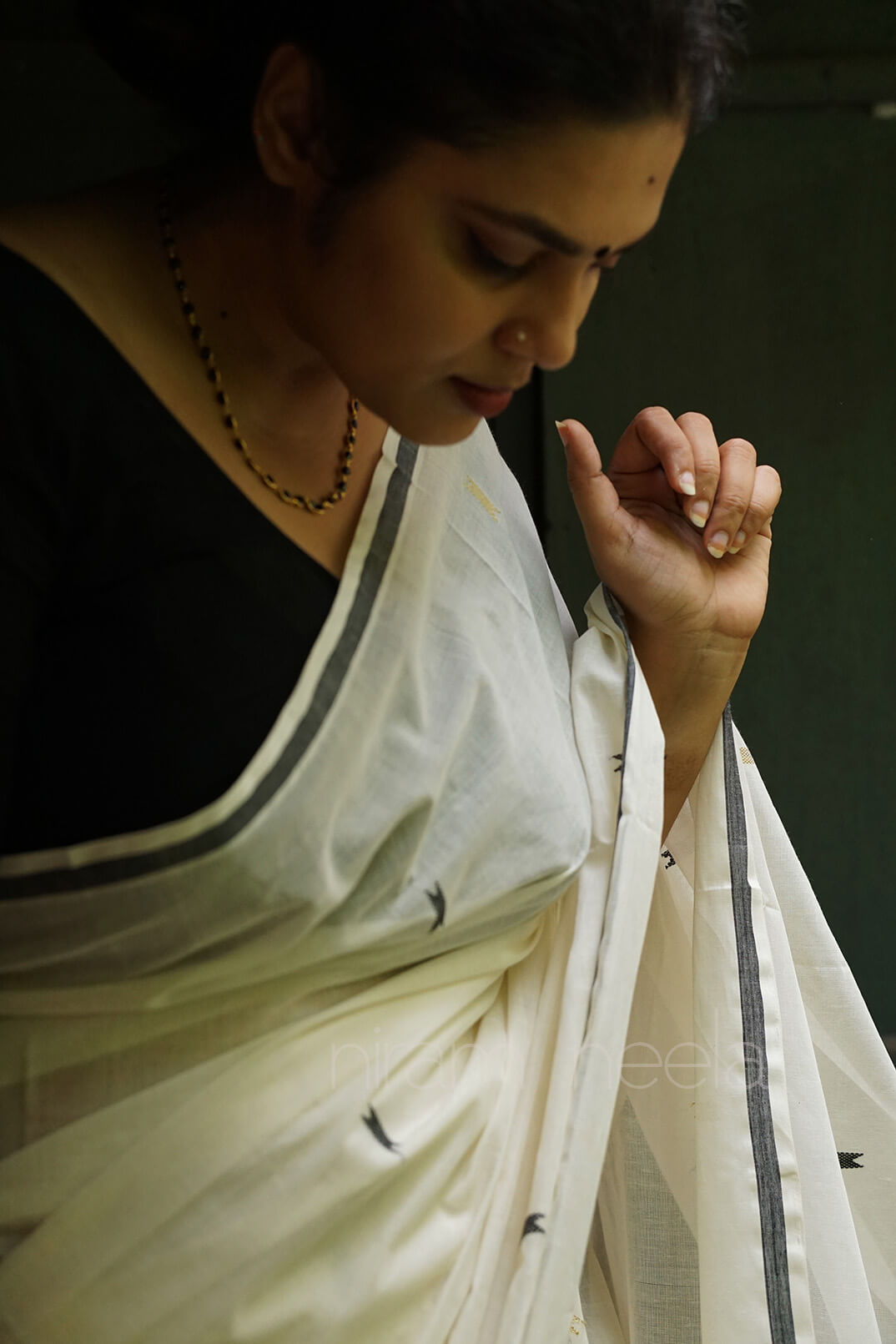 A woman wearing a white sari with black border details and a black blouse, captured in a moody, low-light portrait | Kasavu by Niram Neela