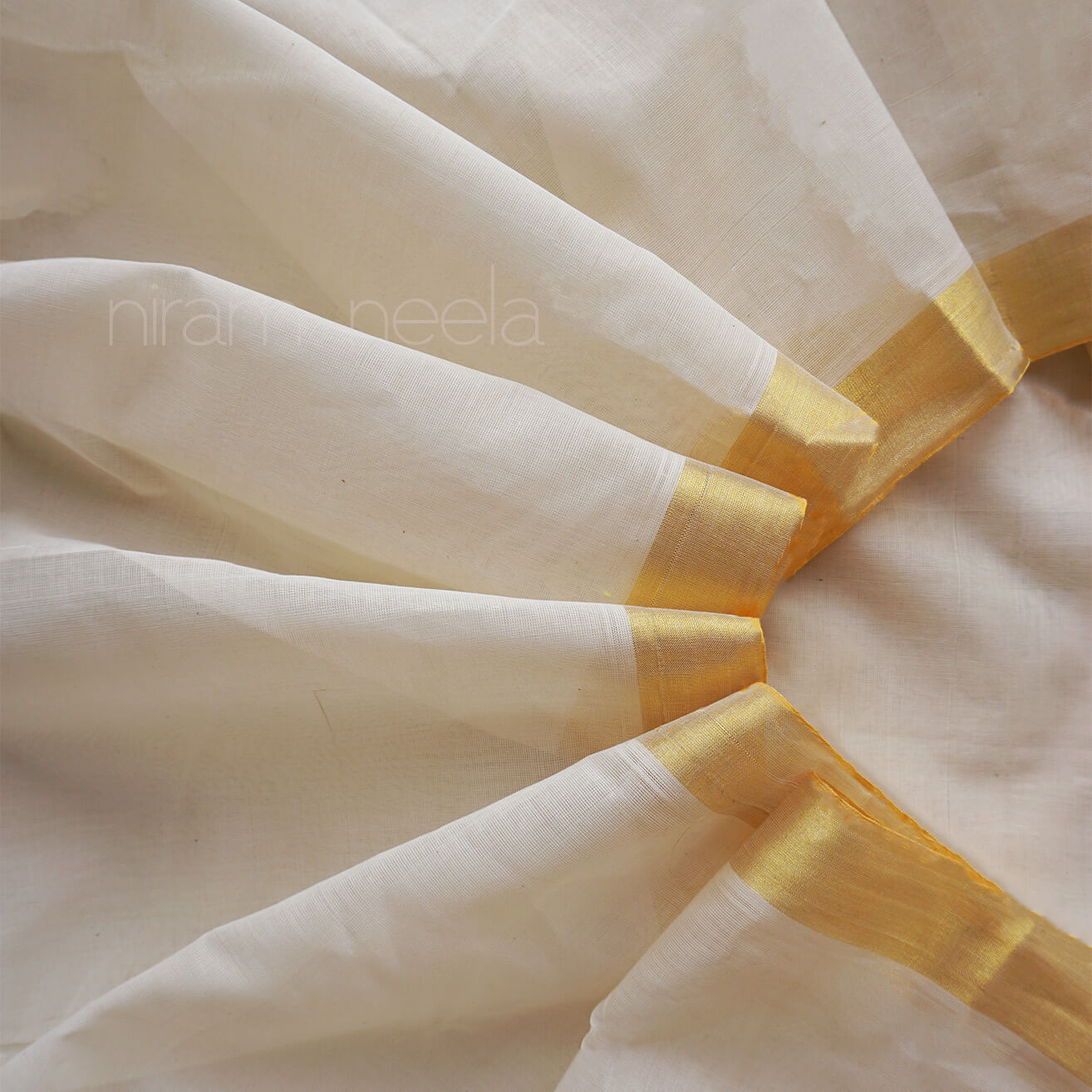 A close-up of draped off-white saree with a shimmering gold border, arranged in soft folds. The watermark 'niram neela' is visible across the center.