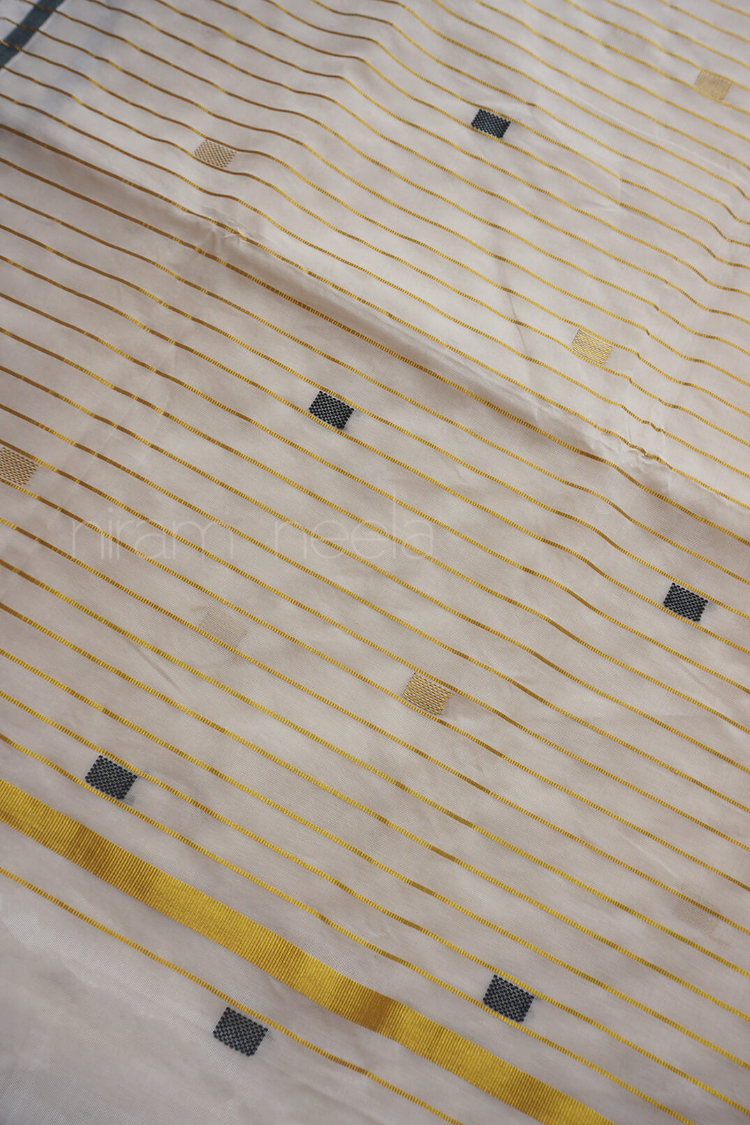 A close-up of a cream-colored fabric with thin horizontal gold stripes and scattered small black rectangular accents.