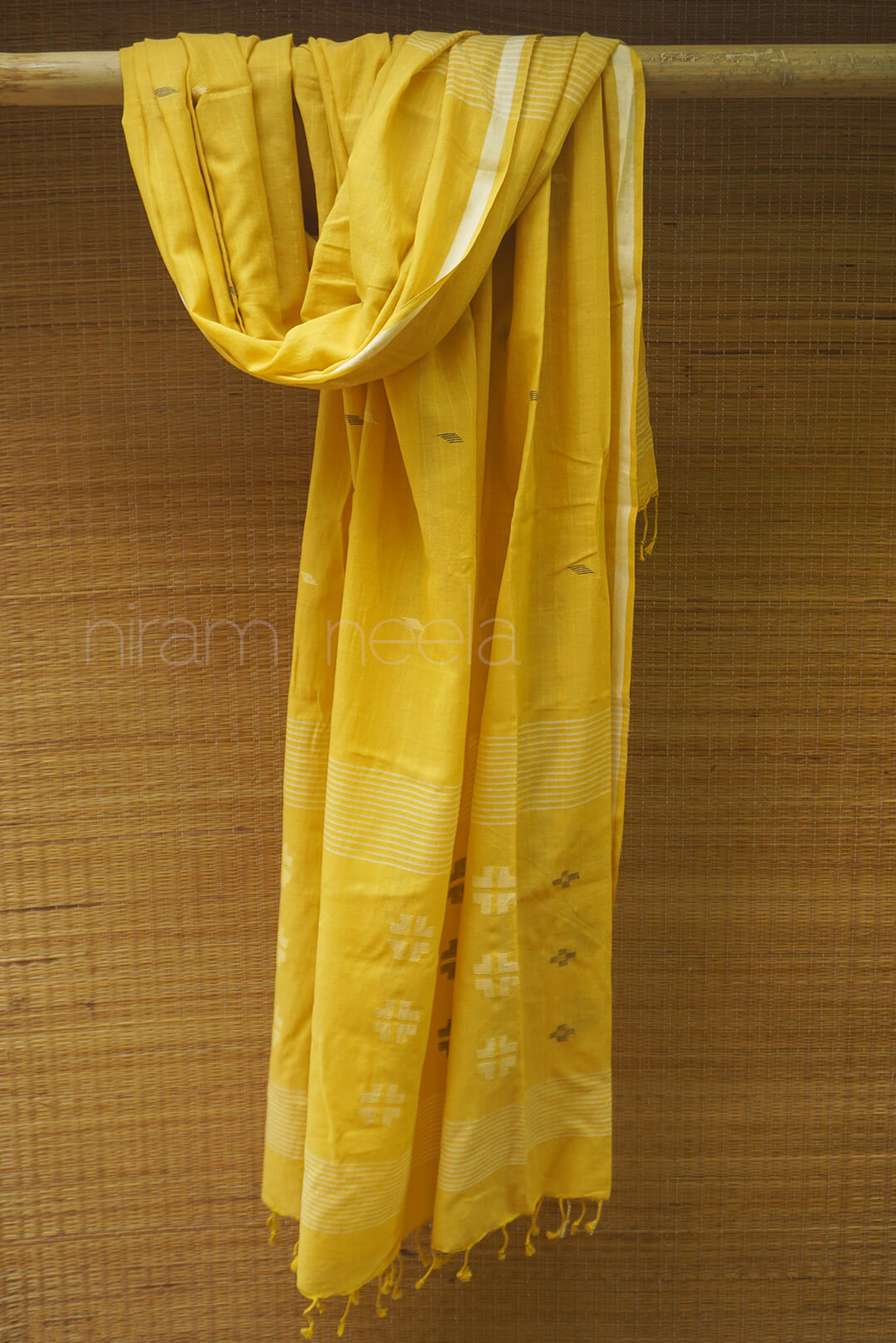 Yellow scarf with white patterns draped over a wooden rod against a textured brown background.