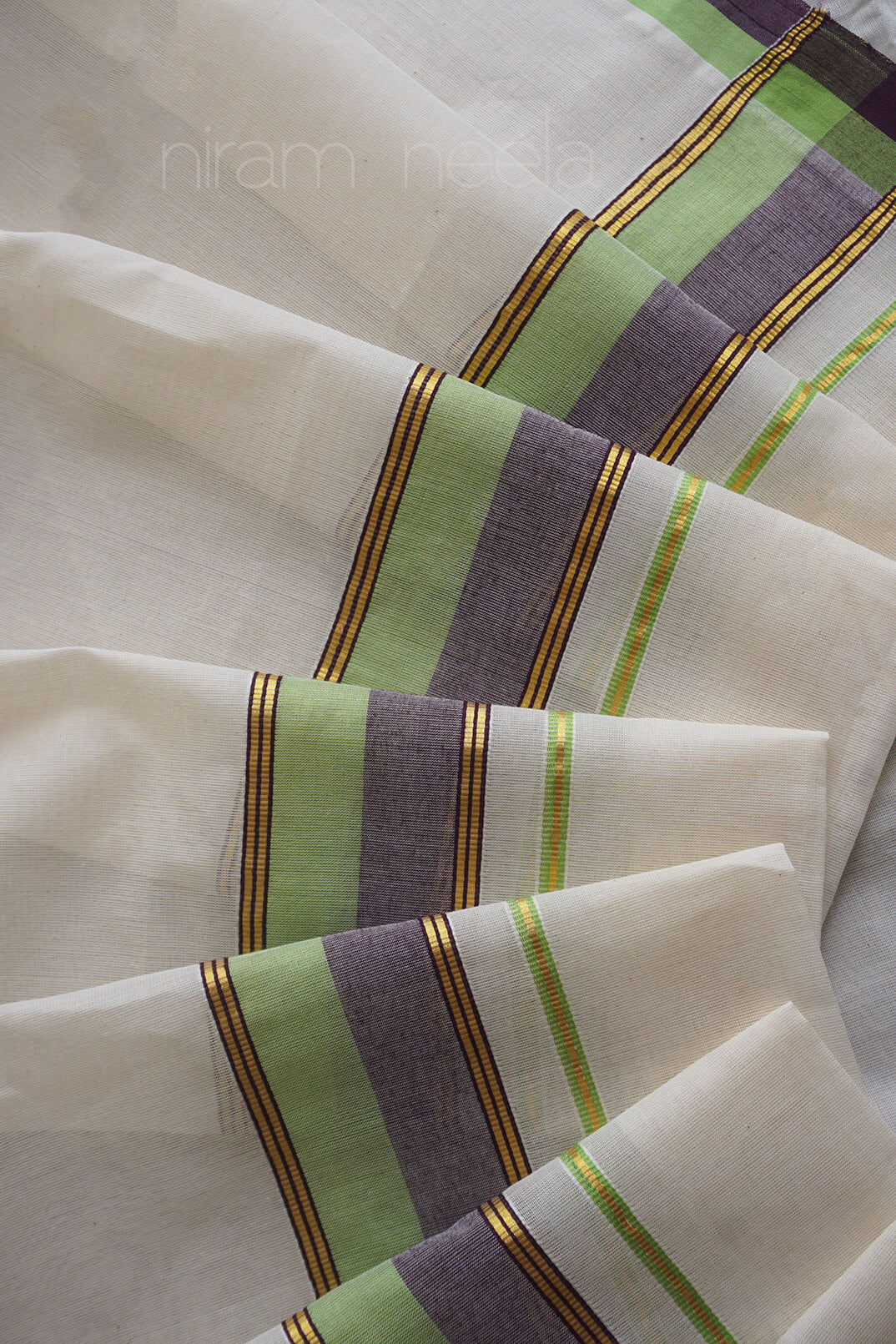 A close-up of an off-white set mundu with stripes in lime green, muted purple, and thin gold lines, arranged in soft folds. A faint 'niram neela' watermark is visible near the top.