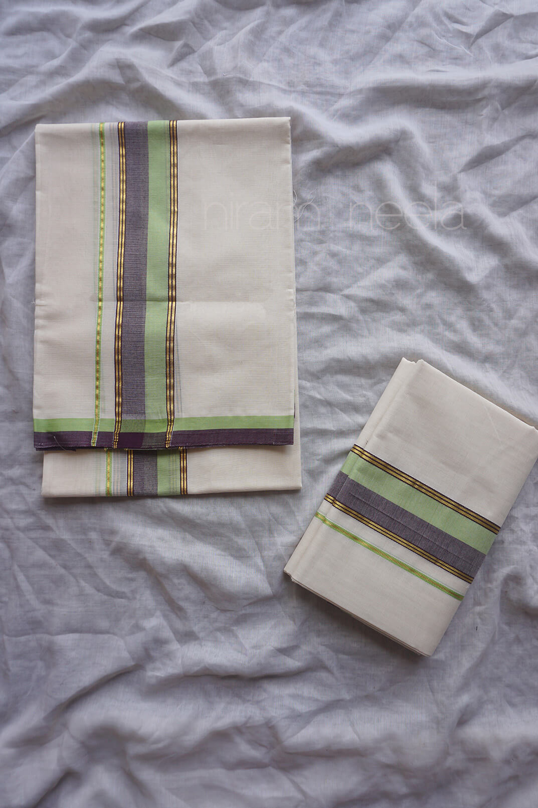 Folded cream-colored cotton set mundu with green, purple, and gold striped borders, arranged on a light gray fabric background.